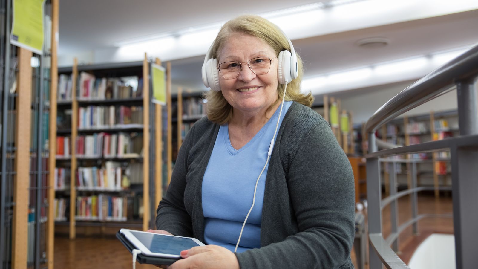 Smiling older woman wearing white headphones and holding a tablet in a library
