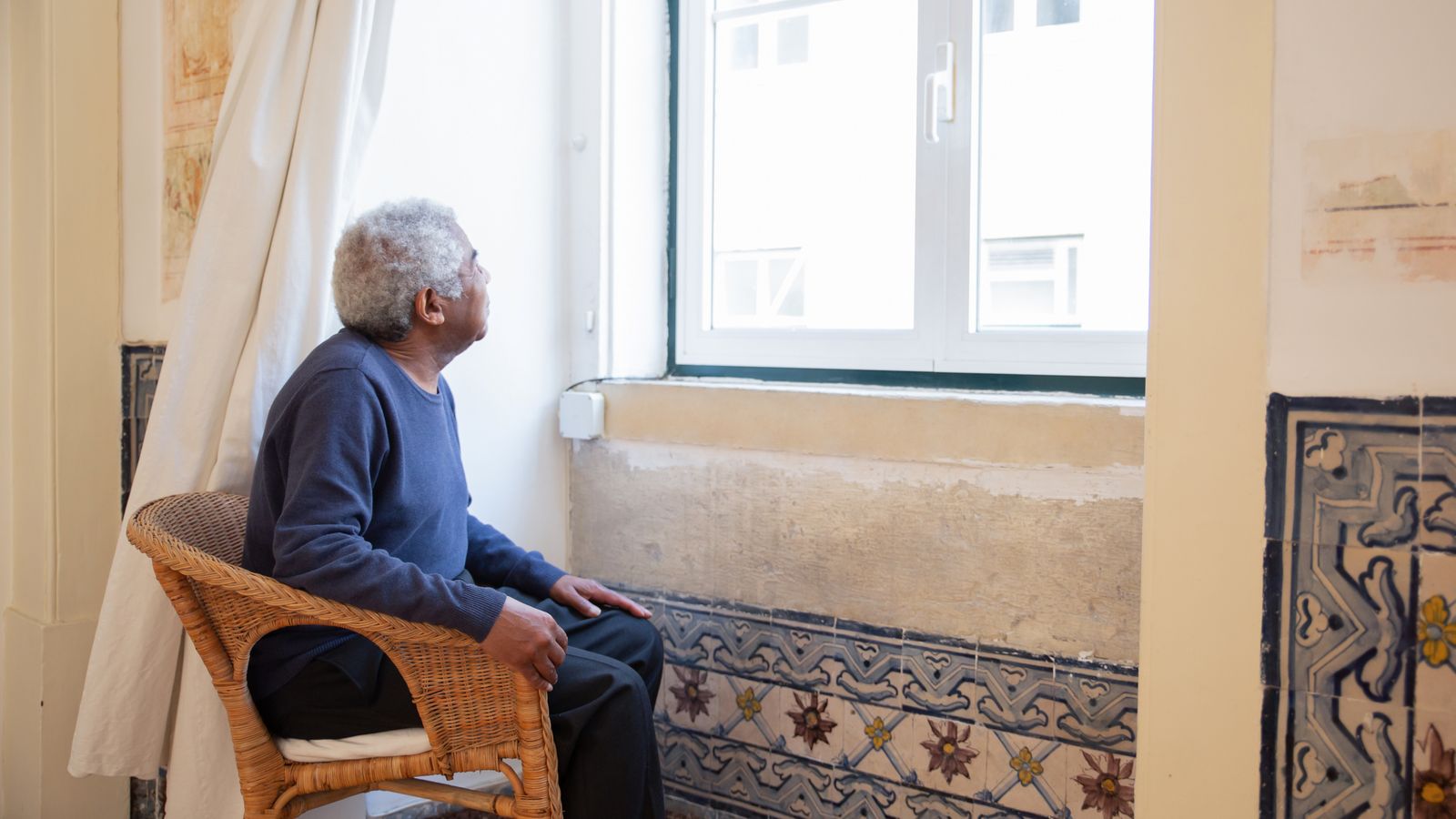 A care worker sits close beside an older man, holding his hand as he looks down, quietly listening