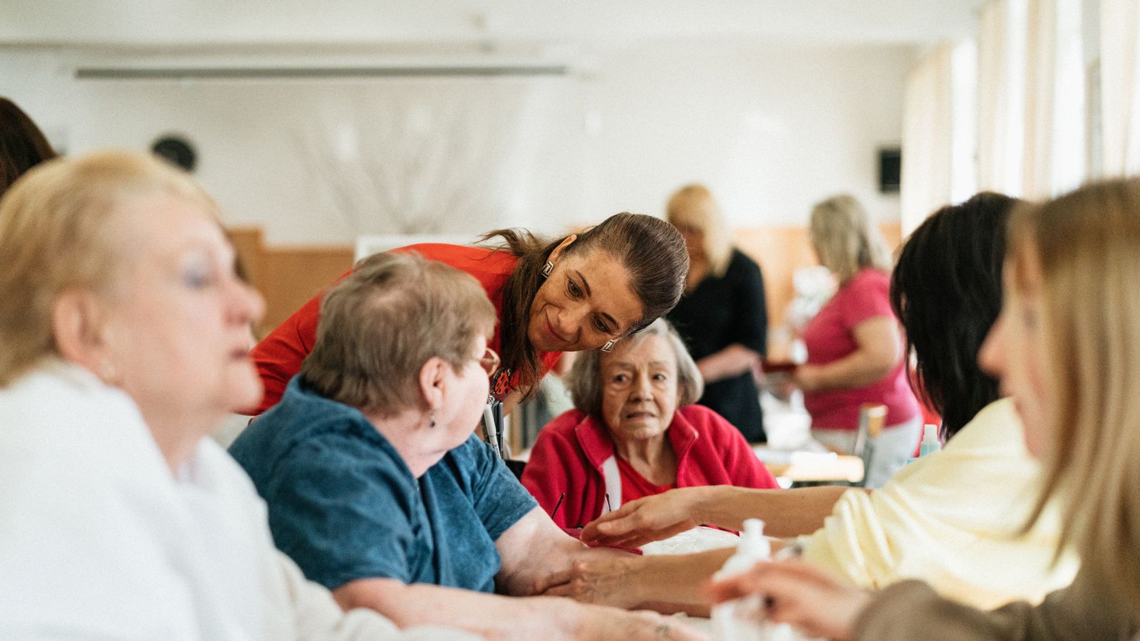 An activity leader leans in to listen to an older woman at a table of seniors during a group session in a bright community room