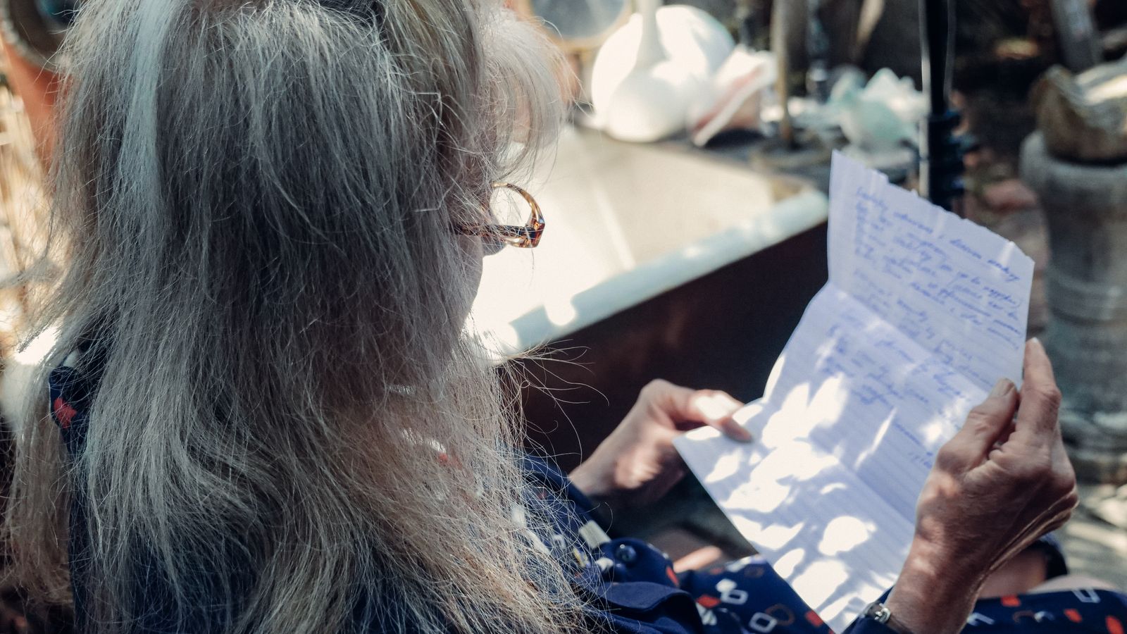 An older man sits by a window with a notebook and old photographs, looking back over his life