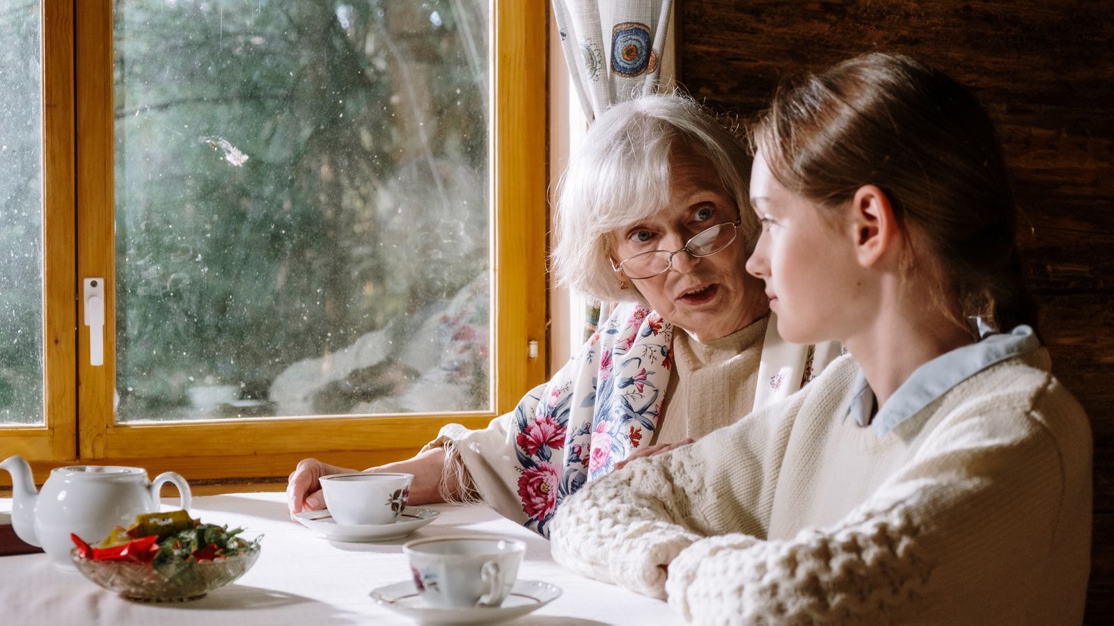 An older woman and a younger companion look through a photo album together at a kitchen table, mid-conversation