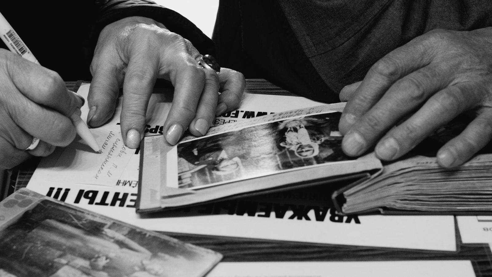 Close-up of older hands turning the pages of a black-and-white photo album while another person writes captions beside it